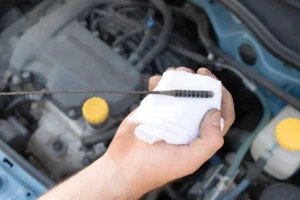 How to Check Oil Level in Car; Realistic close‑up documentary photograph taken from a slightly high‑angled view of an adult light‑skinned male’s right hand holding a dipstick. The hand shows short dark forearm hair, visible knuckles, short fingernails with dirt smudges, and grips a small balled white cotton rag (clean with faint lint). The dipstick is a thin dark metal rod with twisted‑wire texture, a flat elongated tip bearing evenly spaced deep grooves and dark oily residue, resting horizontally on the rag, palm facing the camera. Behind, an out‑of‑focus car engine compartment reveals a dark gray plastic upper cover with rounded contours and bolt heads, a light‑blue painted inner fender panel with a circular opening, two yellow plastic caps (oil fill cap on the cover and cap on a translucent white fluid reservoir), the reservoir containing clear fluid and connected by light‑blue‑green and black hoses, plus assorted black, grey and blue plastic hoses, electrical wires, and silver‑gray metal parts. Lighting is natural diffused from above‑front‑left, creating soft shadows; muted industrial tones of greys, desaturated blues, yellows and whites with moderate contrast; shallow depth of field keeps hand, rag and dipstick sharp while the engine background is blurred, giving an utilitarian, informative tone.